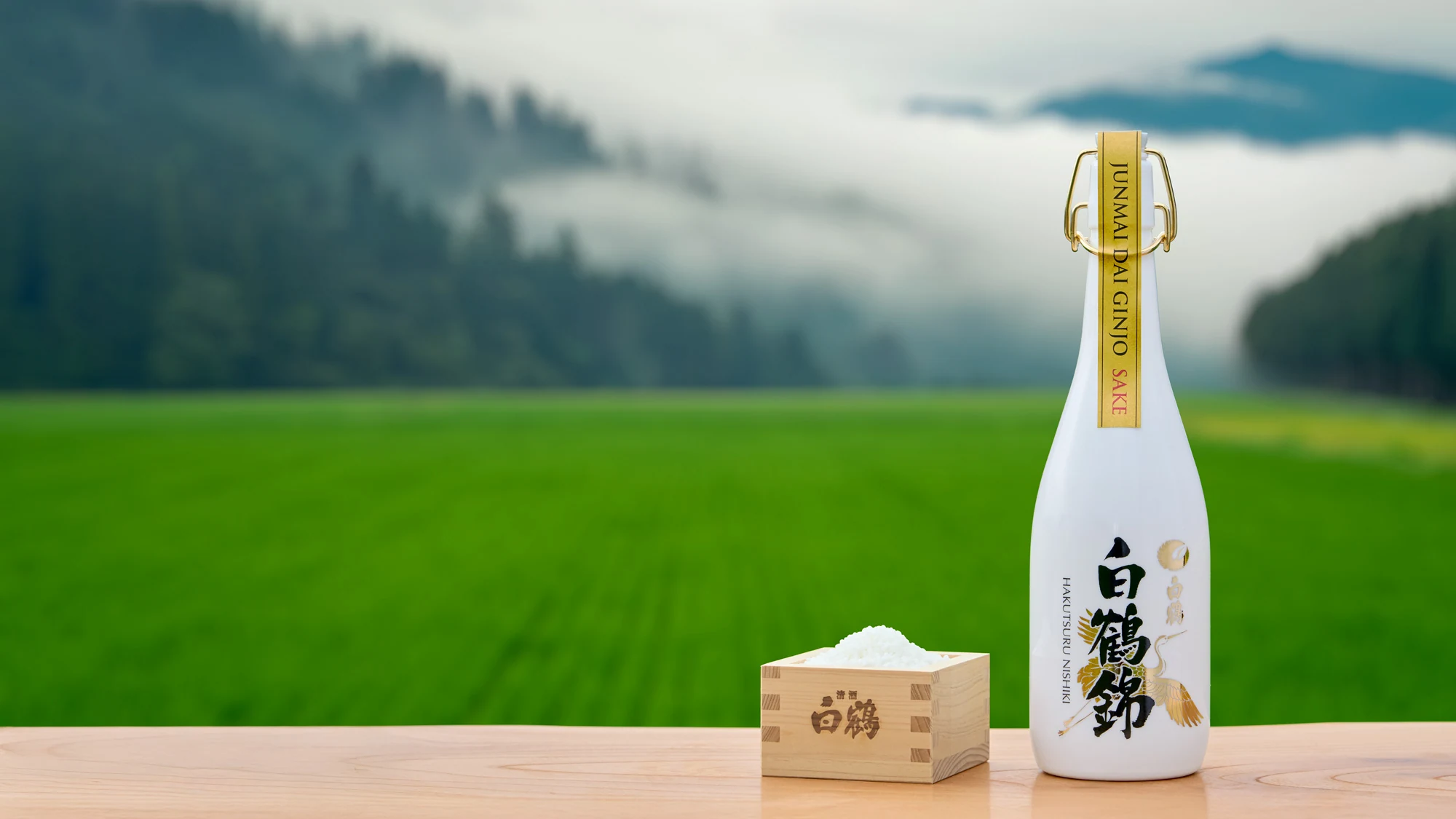 A Hakutsuru sake bottle and masu set against green rice fields and misty mountains