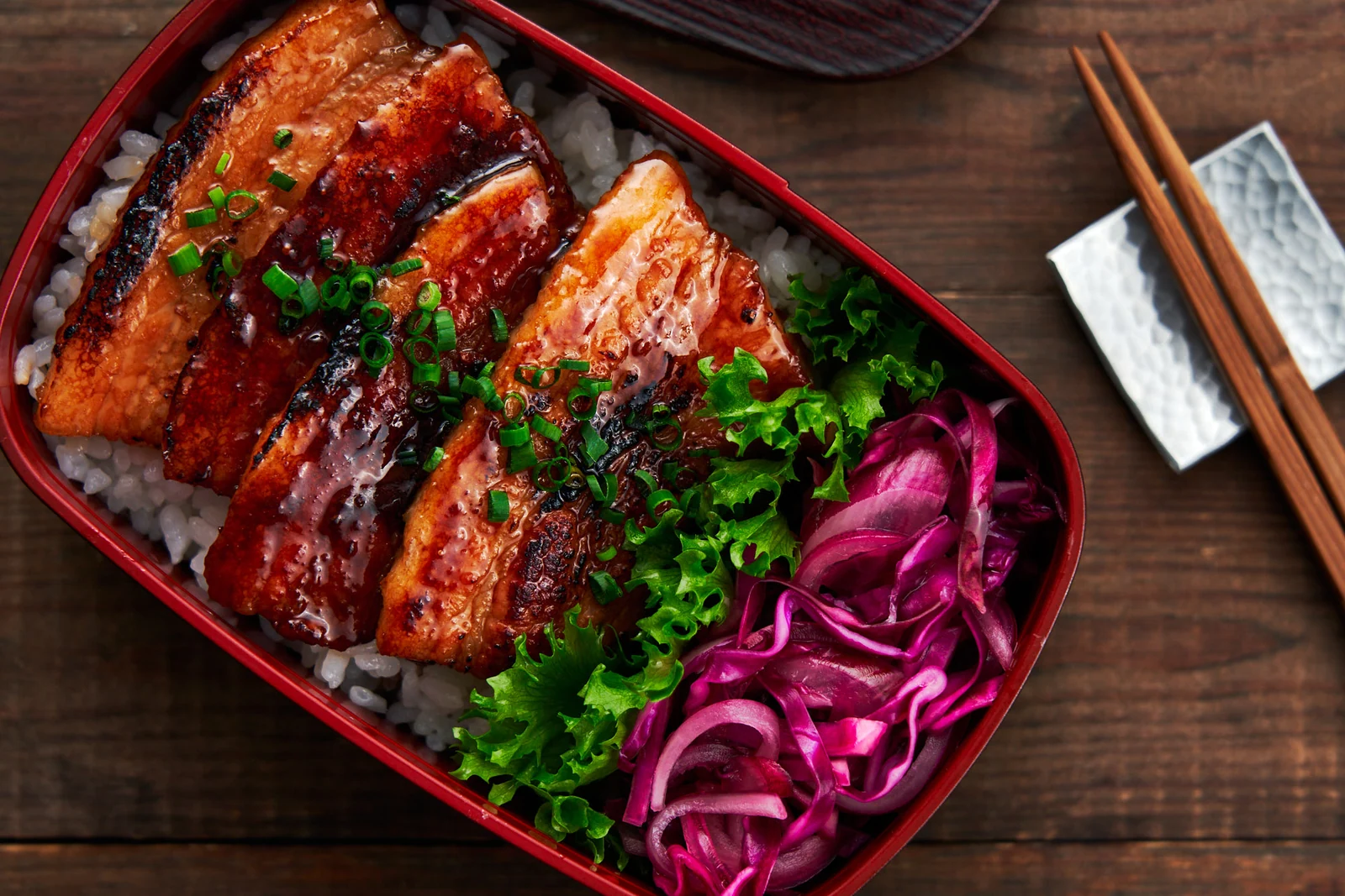 Glazed slices of pork arranged over rice with pickled cabbage in a bento box