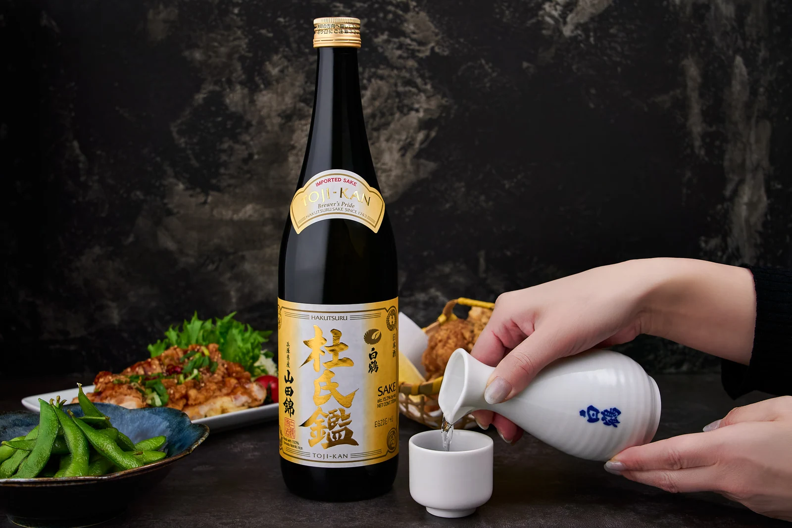Sake being poured beside a Hakutsuru bottle and a spread of Japanese dishes