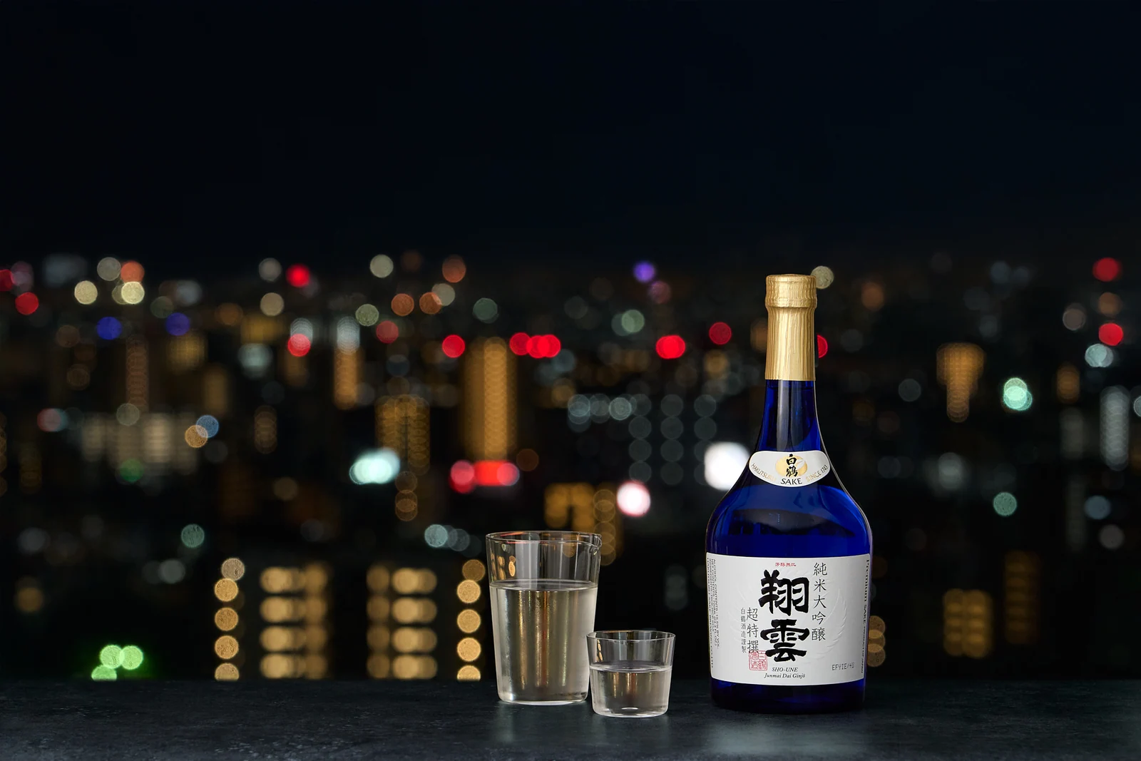 A bottle and glass of Hakutsuru Sho-Une sake against city lights at night
