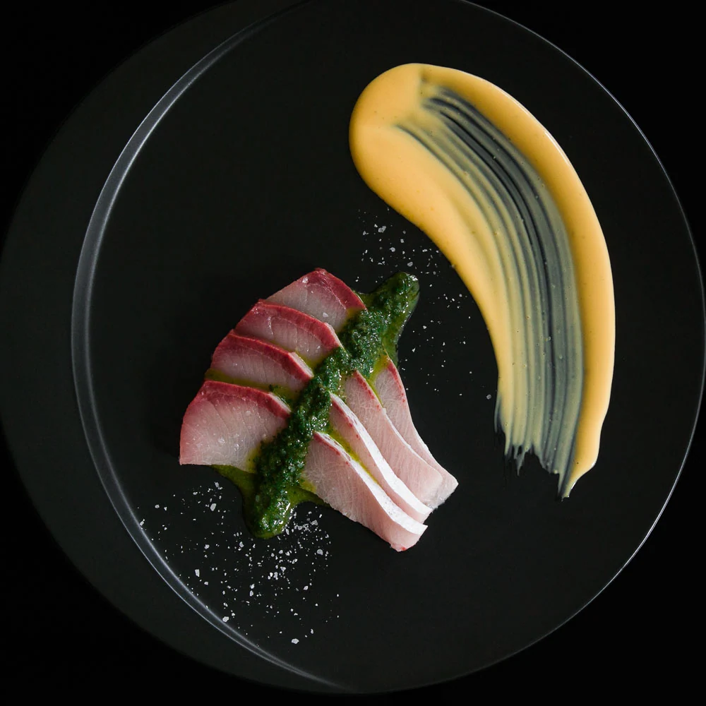 Slices of yellowtail arranged with vegetables and a golden sauce on a black plate